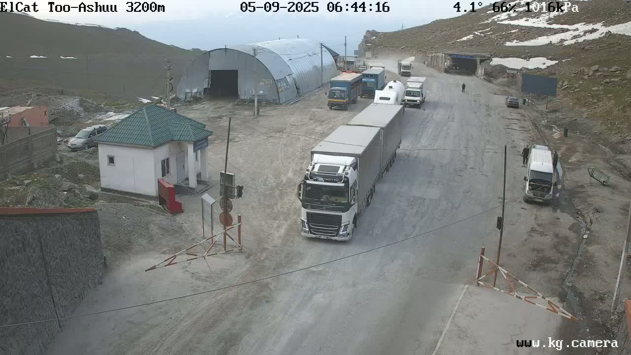 A mountain road with several trucks and cars, a small building, and a large metal structure, under partly cloudy skies.