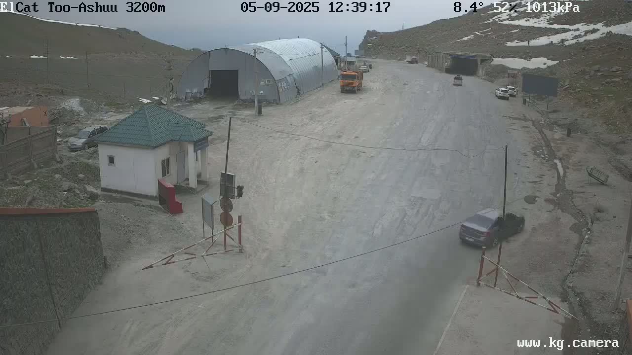 A mountain road with a few vehicles, a small building, and a large metal structure under an overcast sky.