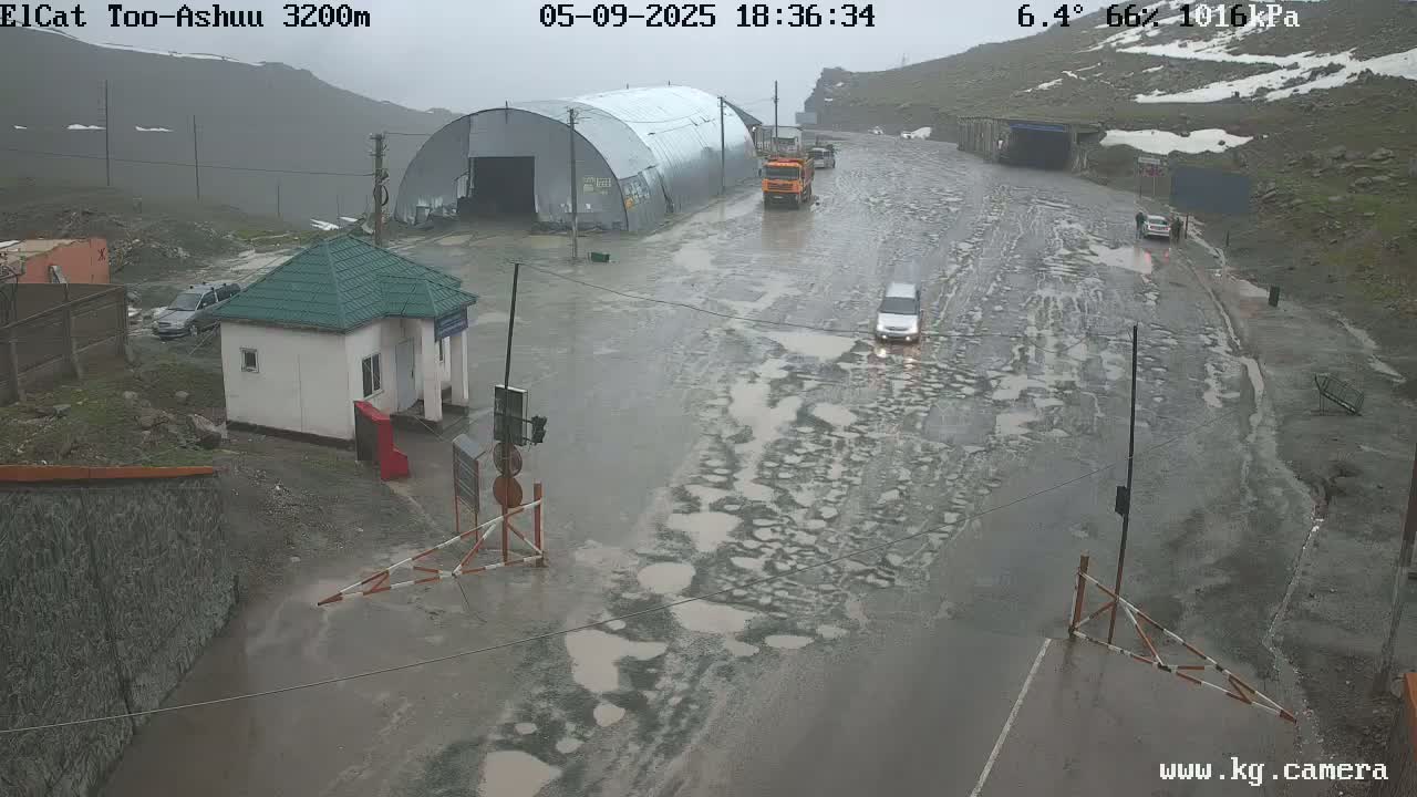 A wet, pothole-ridden road at a mountain pass checkpoint, with a small building and several vehicles present in rainy weather.