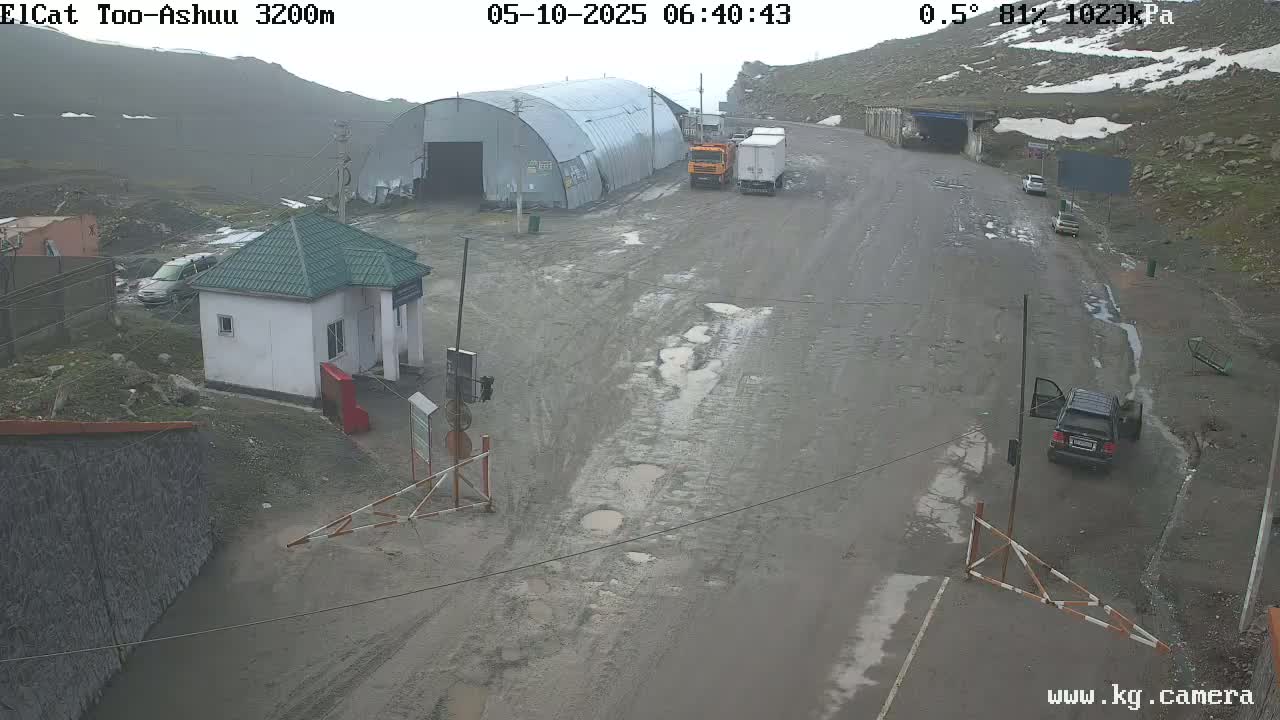 A mountain road with several vehicles, a small building, and a large metal structure is seen on an overcast day.