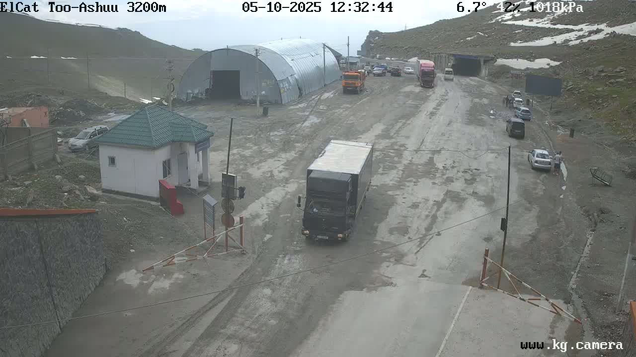 A wet, overcast mountain road with several vehicles, including trucks and cars, near a small building and a large metal structure.