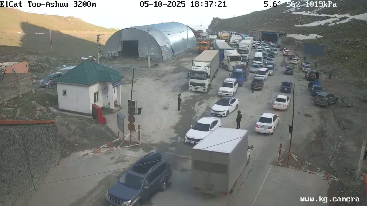 A long line of vehicles waits at a mountain pass border crossing under a slightly overcast sky.
