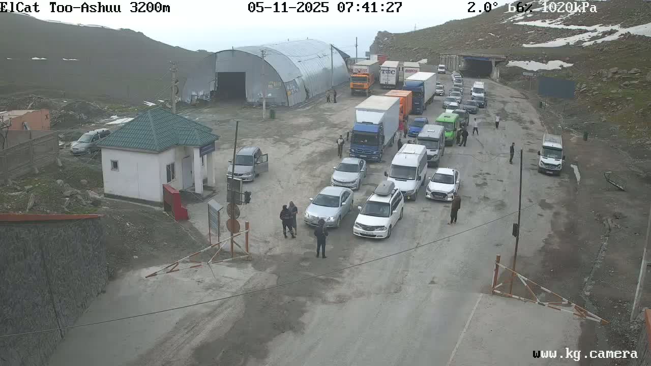 A line of vehicles waits at a mountain pass border crossing under overcast skies.