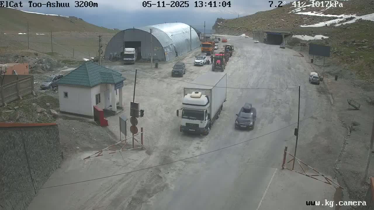 A mountain road with several vehicles, including trucks and cars, near a small building and a large metal structure, under a cloudy sky.