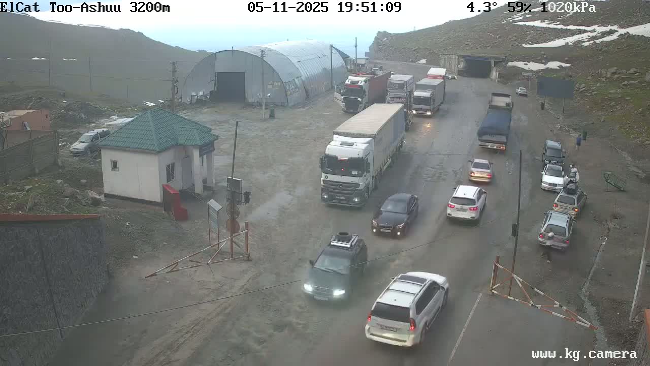 A wet road with numerous vehicles, including several trucks, cars, and SUVs, is seen near a small building and a larger metal structure in a mountainous area under overcast skies.