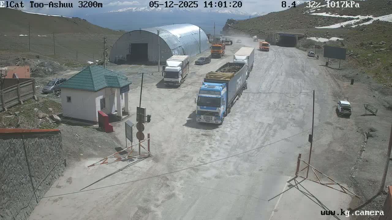 A mountain road with several trucks and cars, a small building, and a large metal structure is visible under a partly sunny sky.