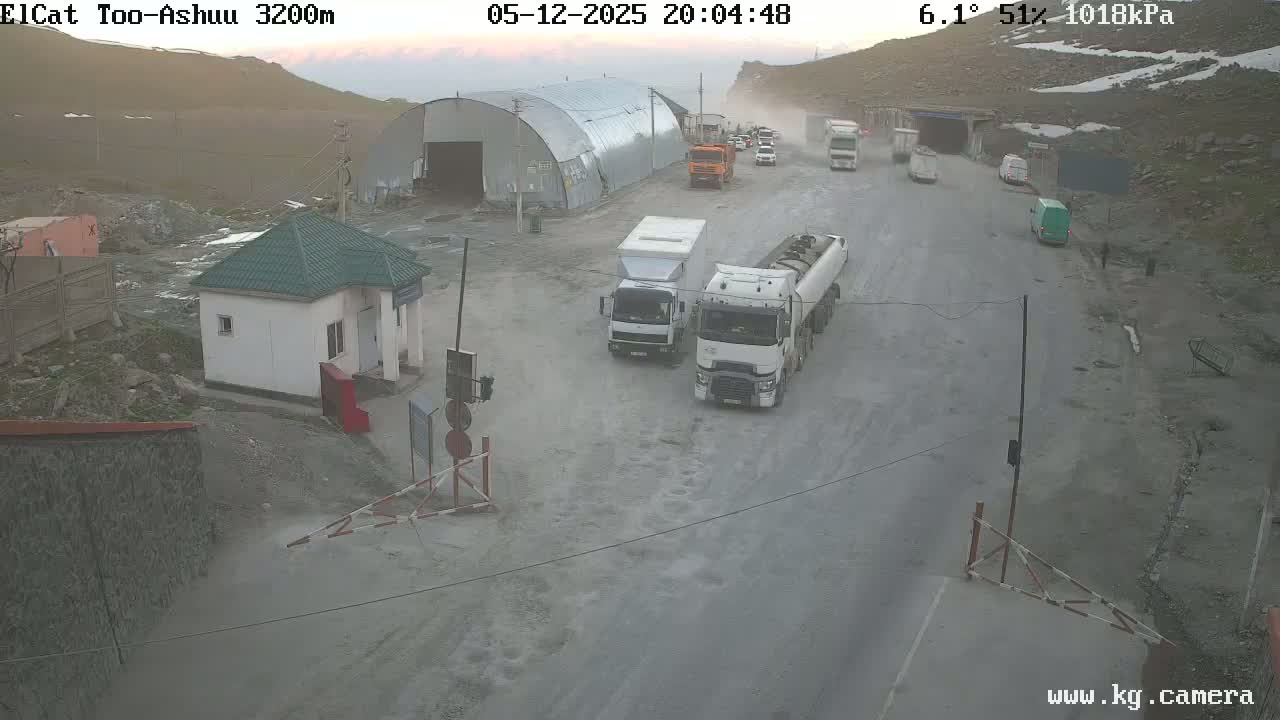A mountain road with several trucks and a small building, with dusty conditions and a partly cloudy sky.
