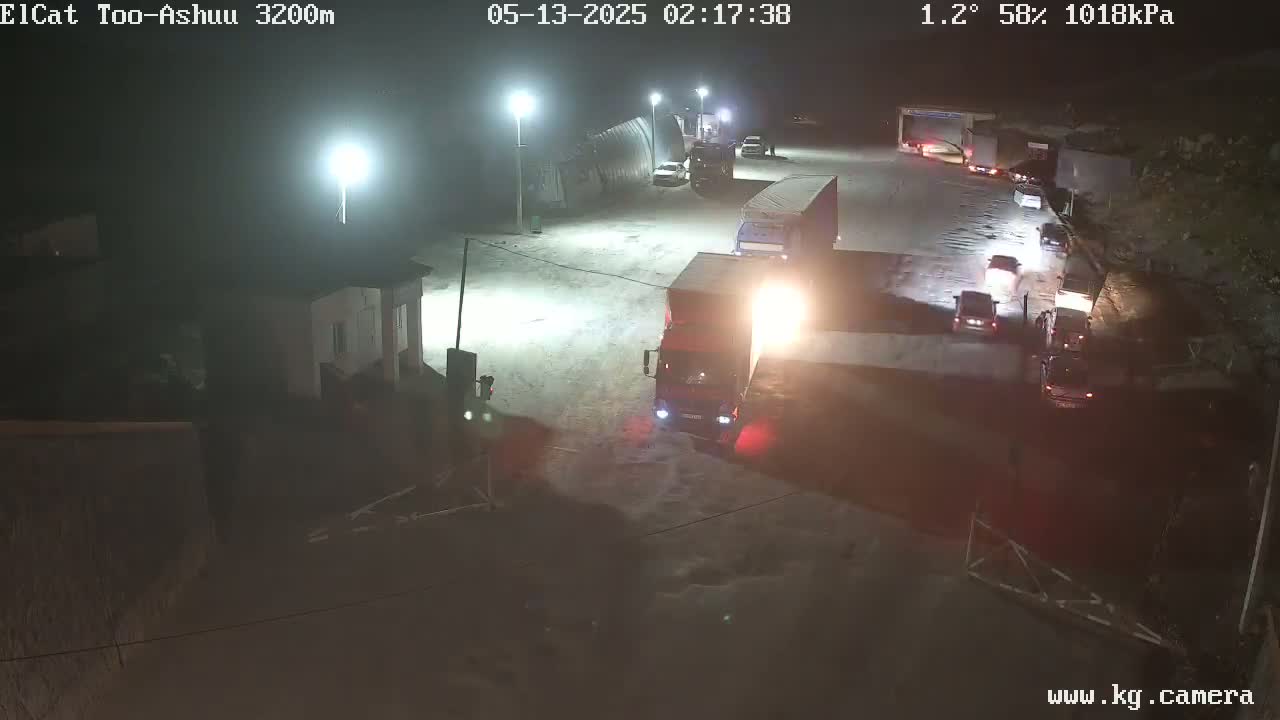 At night, under a mostly clear sky, several vehicles, including trucks and cars, are parked and moving in a large, well-lit area.