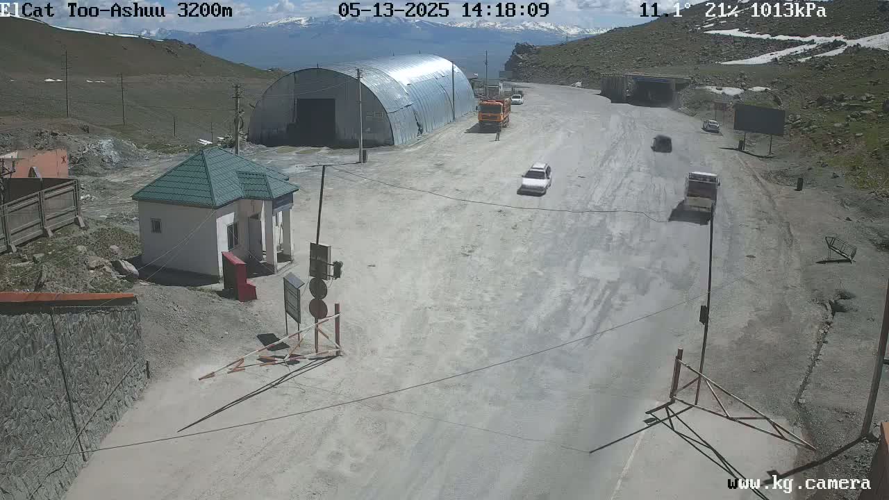 A high-angle, sunny-day view of a mountain road with several vehicles near a small building and a larger metal structure, all on a dusty, light-gray surface.