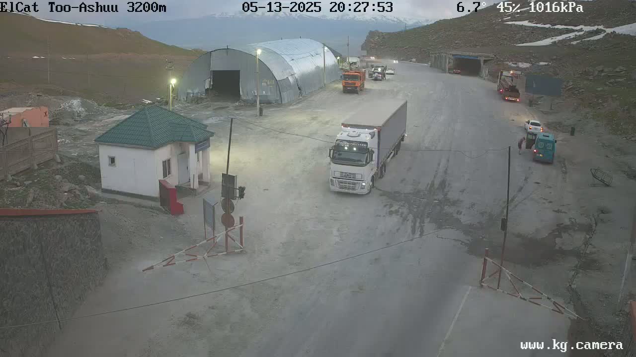A mountain pass border crossing with several vehicles, a small building, and a large metal structure under slightly overcast conditions.