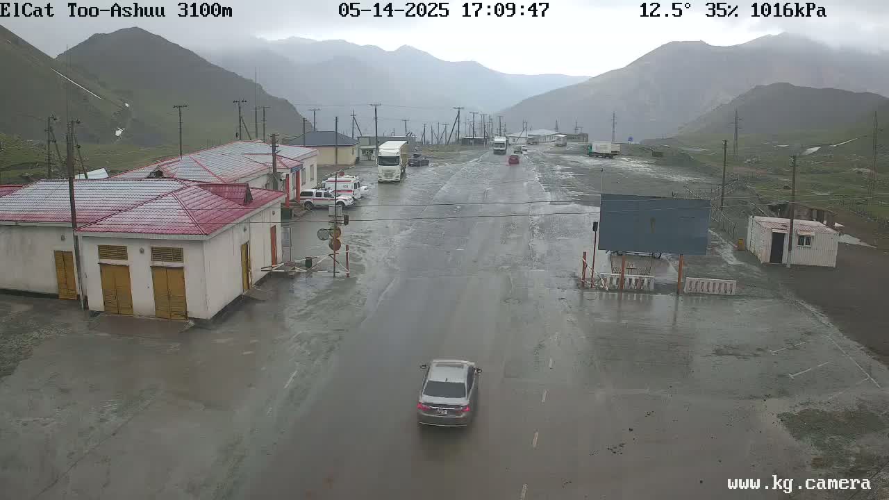 A flooded roadway in a mountainous area with several buildings and vehicles, under overcast skies.