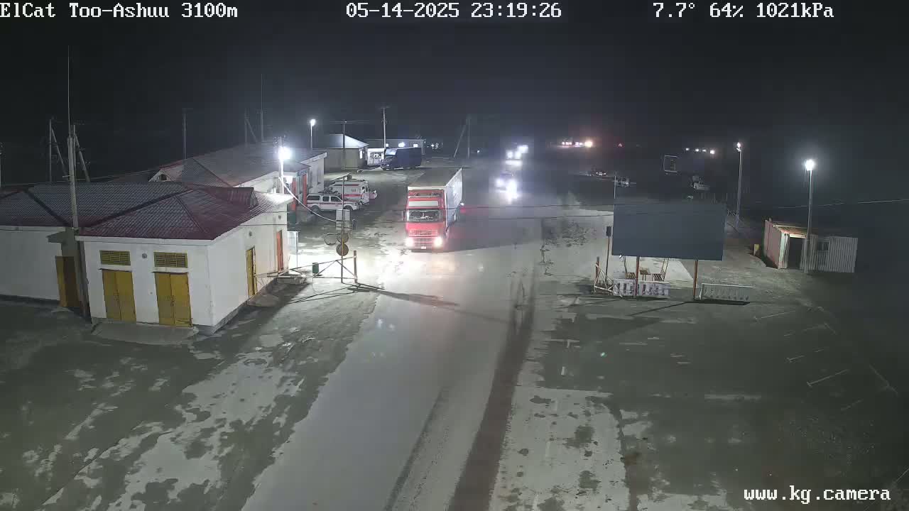 A nighttime view of a roadway and buildings, with a red semi-truck and other vehicles parked nearby, under partly cloudy skies at 7.7°C and 64% humidity.