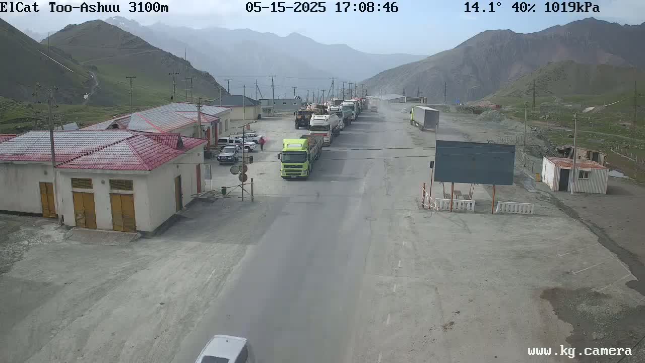 A line of trucks is parked on a roadway in front of a small building complex, situated in a mountainous area under partly cloudy skies, with a temperature of 14.1 degrees and 40% humidity.