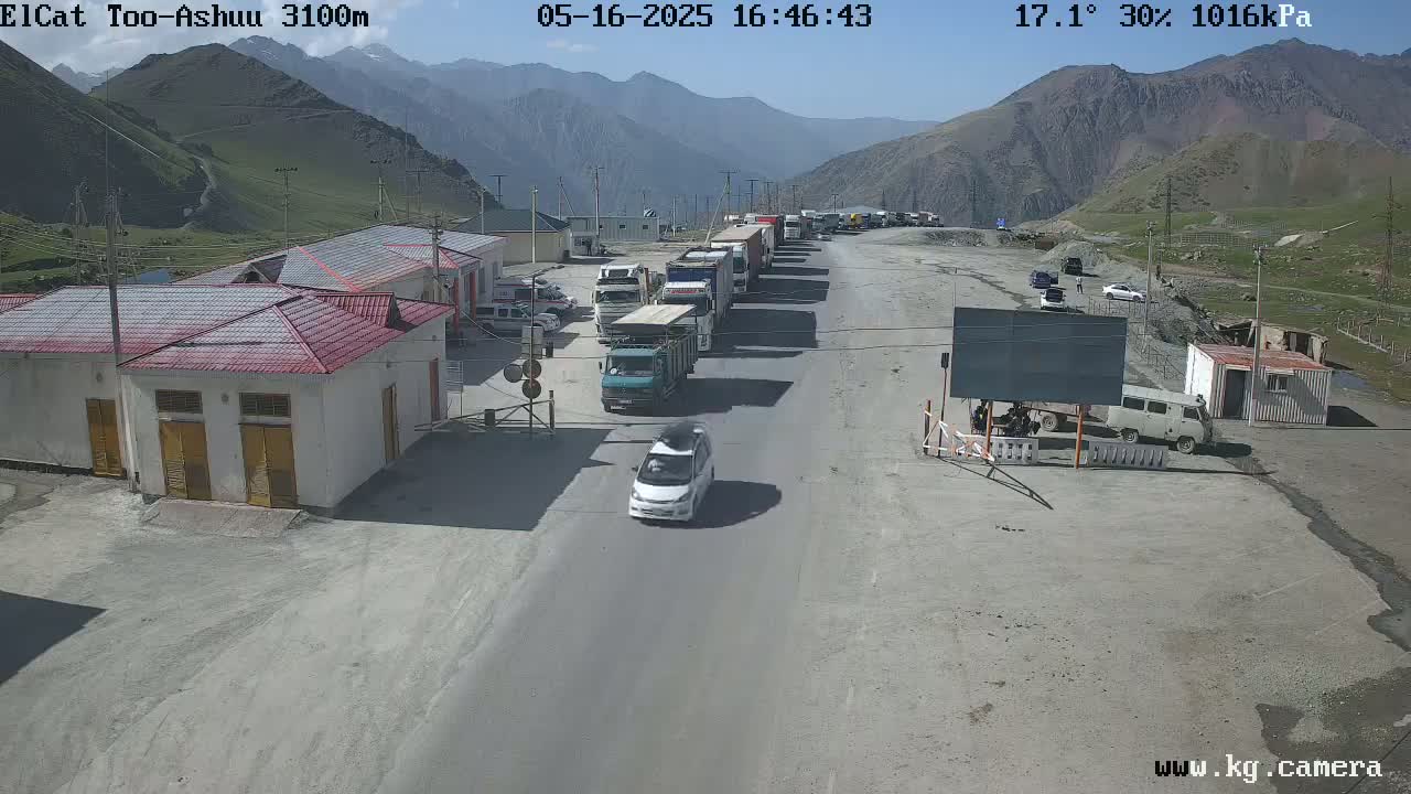 A line of trucks and several cars are parked at a border crossing in a mountainous area under sunny skies.