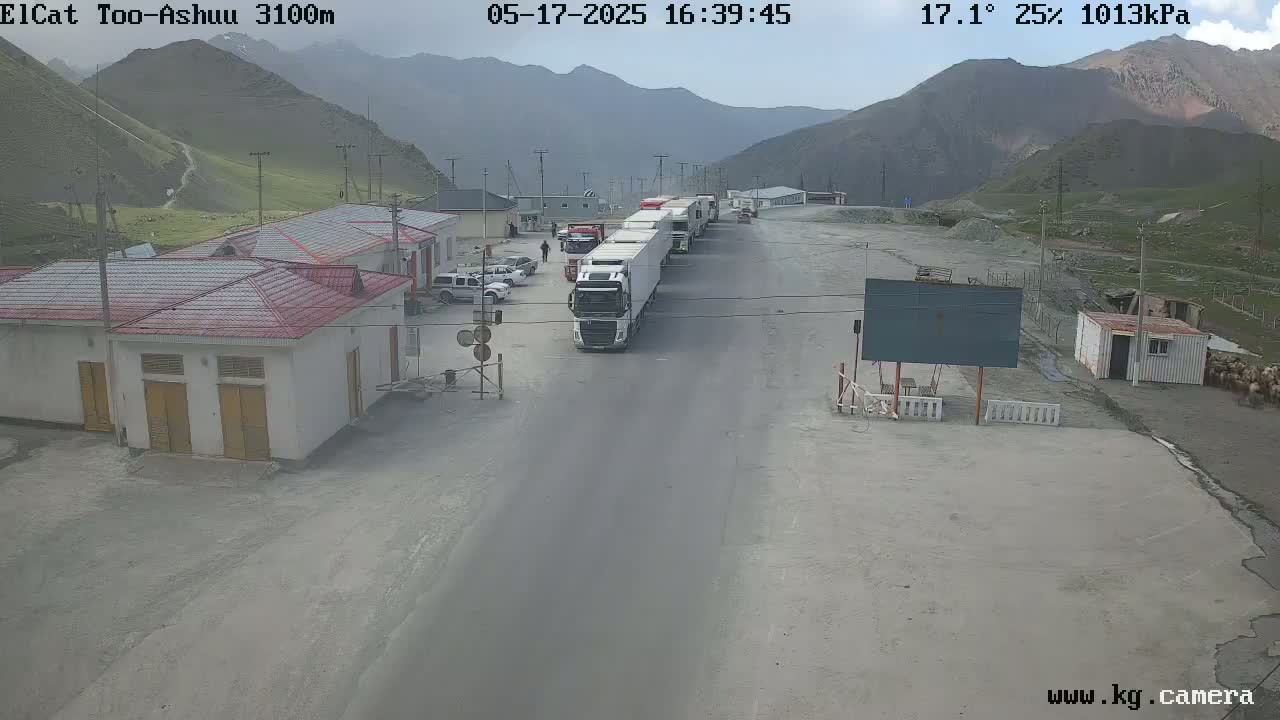 A line of trucks is parked at a border crossing in a mountainous area with partly cloudy skies and a temperature of 17.1 degrees Celsius.