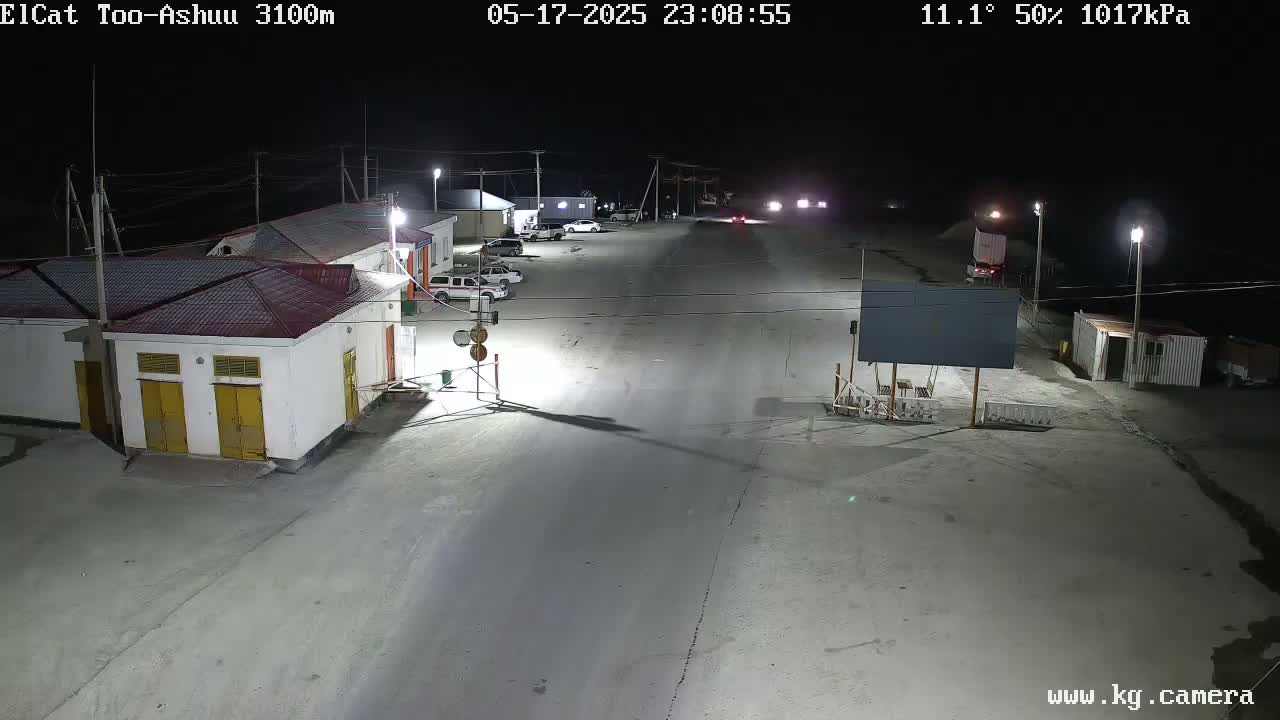 A nighttime, illuminated view of a sparsely populated roadway and several buildings, with a few vehicles parked and some driving down the road.