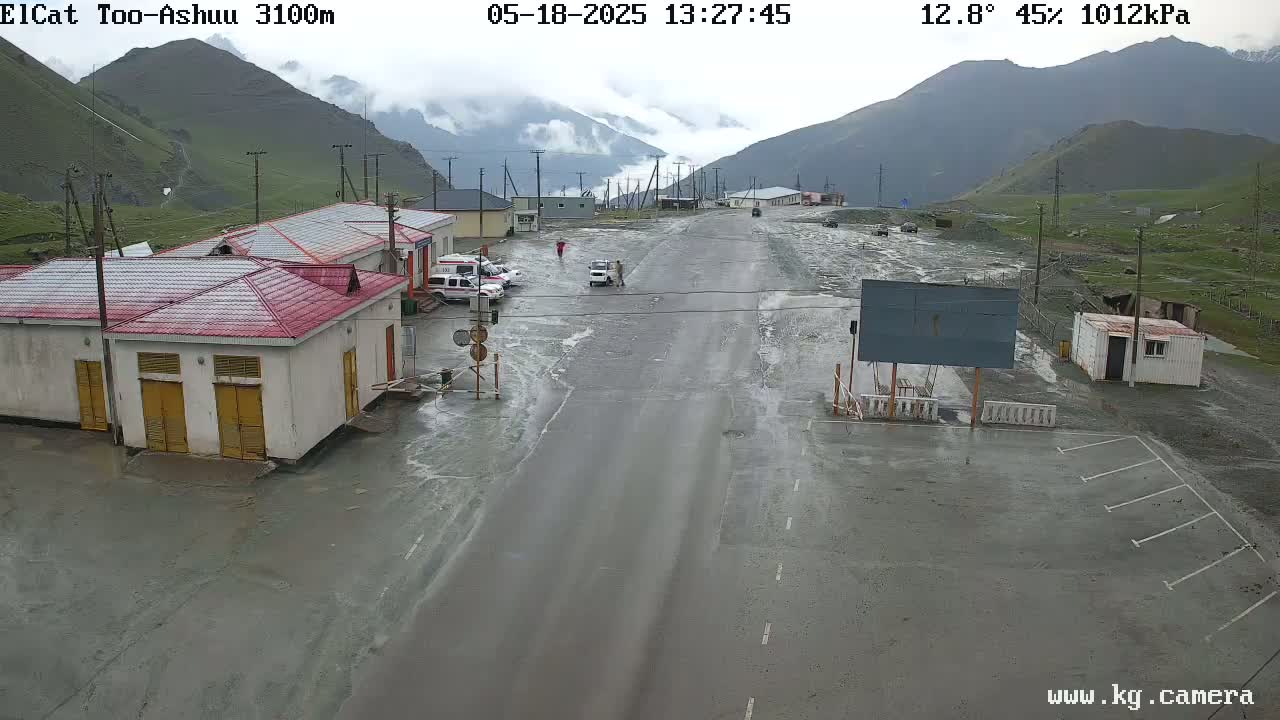 A flooded roadway in a mountainous area passes by several small buildings under an overcast sky.