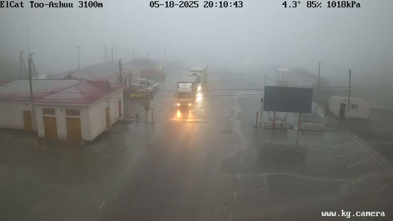 A wet, foggy road with several trucks and cars is seen near a building complex under heavy rain.