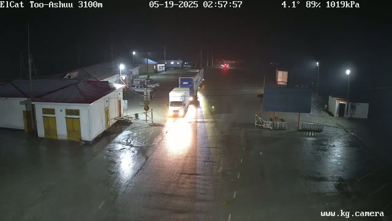 At night in rainy weather, several vehicles, including trucks and cars, are parked and driving on a wet road near buildings.