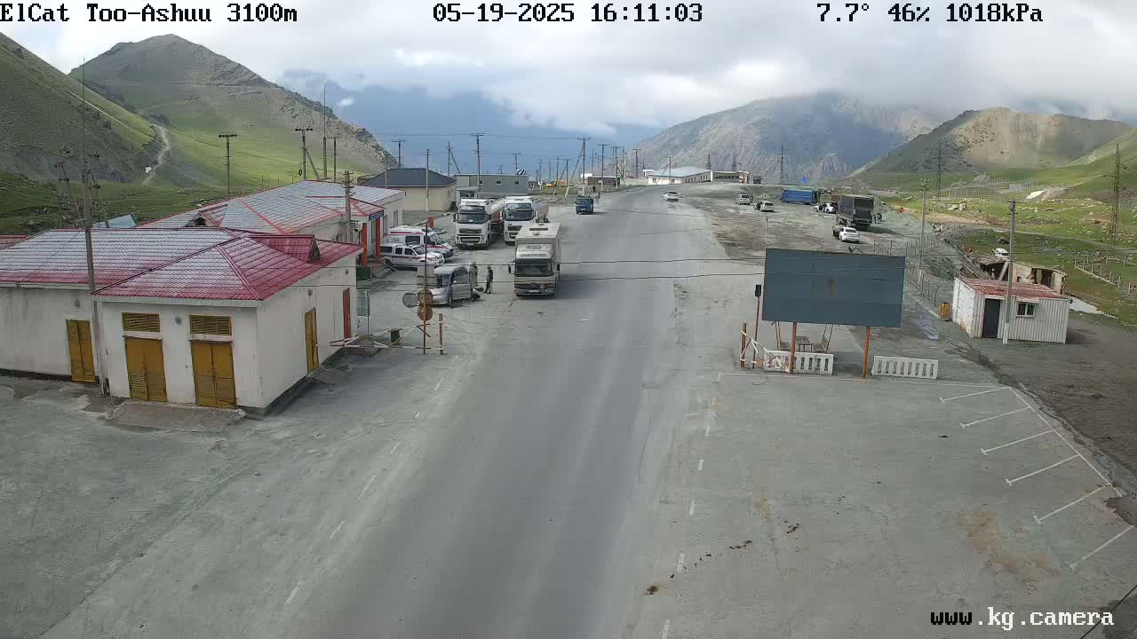 A highway checkpoint in mountainous terrain under partly cloudy skies shows several vehicles parked near small buildings.