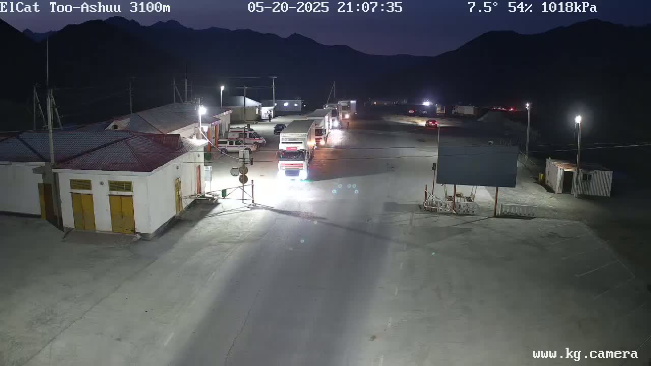 A nighttime, outdoor view of a border crossing with several vehicles, including trucks, parked and driving on a paved area, with buildings and mountains in the background under a clear sky.