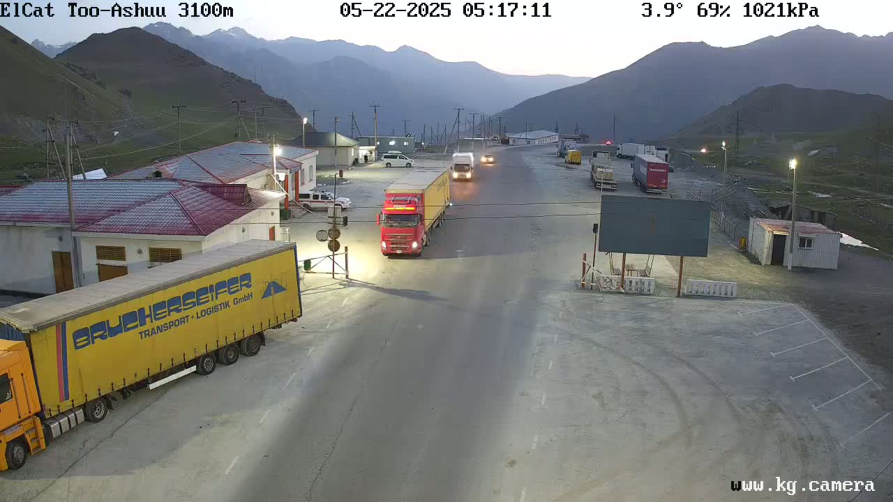 Several large trucks are parked and driving on a road at a mountain pass border crossing at dawn in cool, partly cloudy conditions.