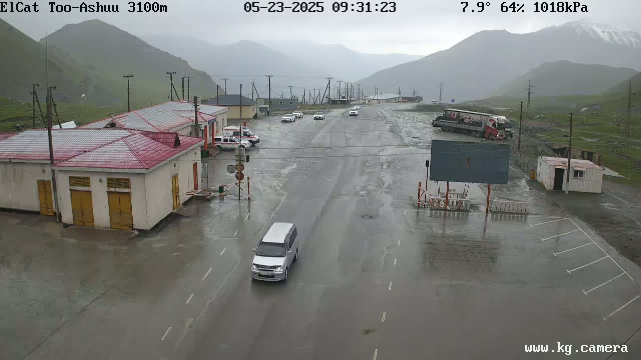 A wet road at a mountain pass checkpoint with several buildings, a few vehicles, and a large truck, under a cloudy, rainy sky.