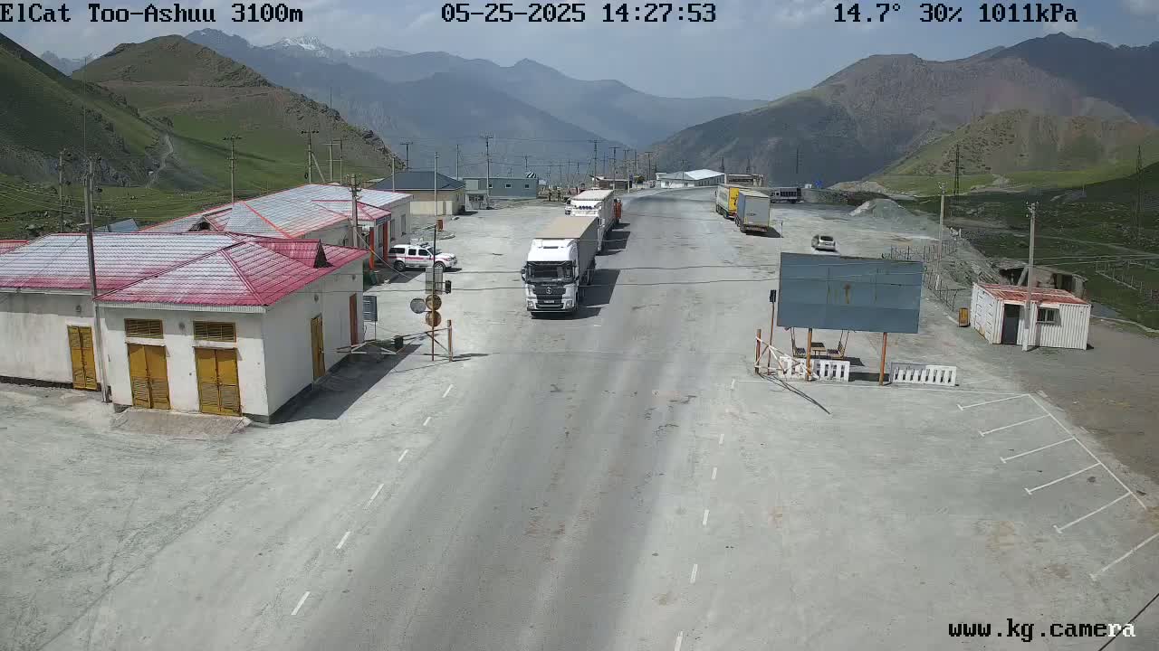 A high-angle, sunny view shows a roadway at a border crossing in a mountainous region, with several buildings, trucks, and cars present.