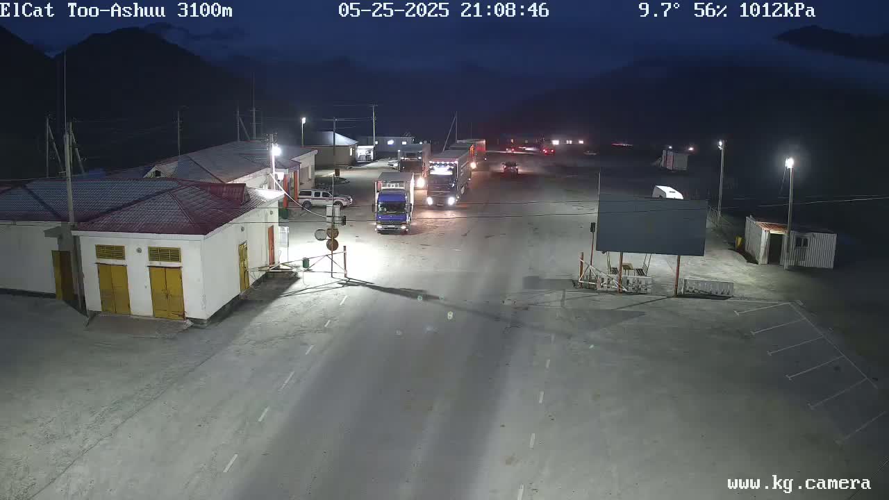 At night, under a 9.7°C, 56% humidity sky, several trucks and cars are parked and driving on a road in front of buildings, in what appears to be a border crossing or checkpoint.