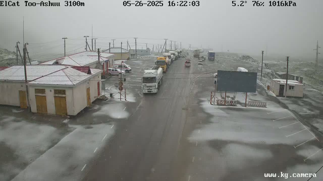 A snow-covered road with several vehicles, including trucks and cars, at a border crossing, with snow lightly dusting the ground and buildings.