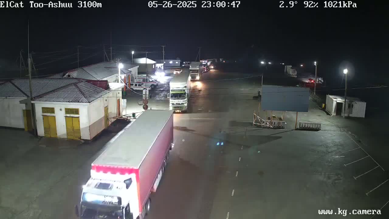 At night, under a clear sky with a temperature of 2.9°C, several large trucks are parked and moving in a lot next to some buildings with snow on the roofs.