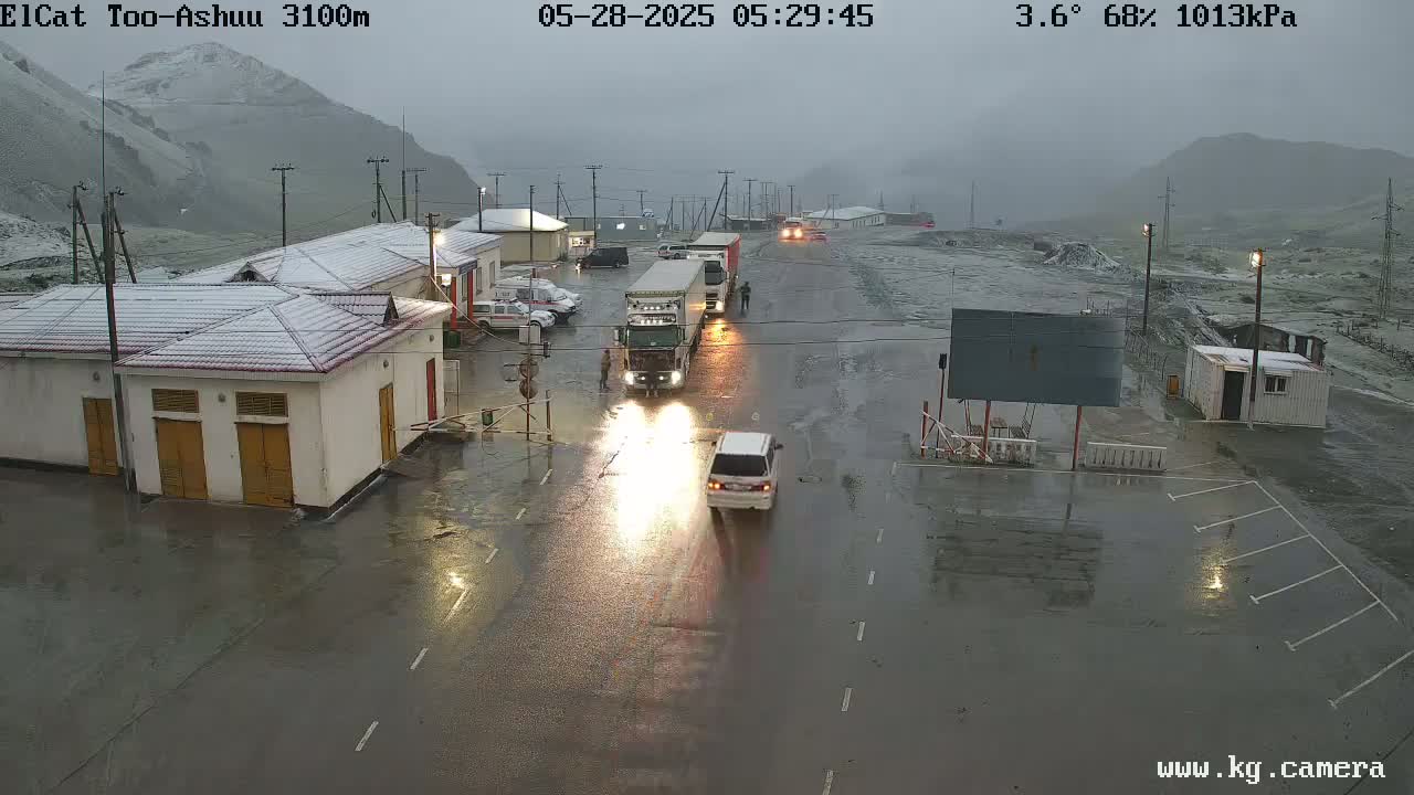 A wet road with several vehicles, including a large truck, in front of buildings covered in snow under an overcast sky.