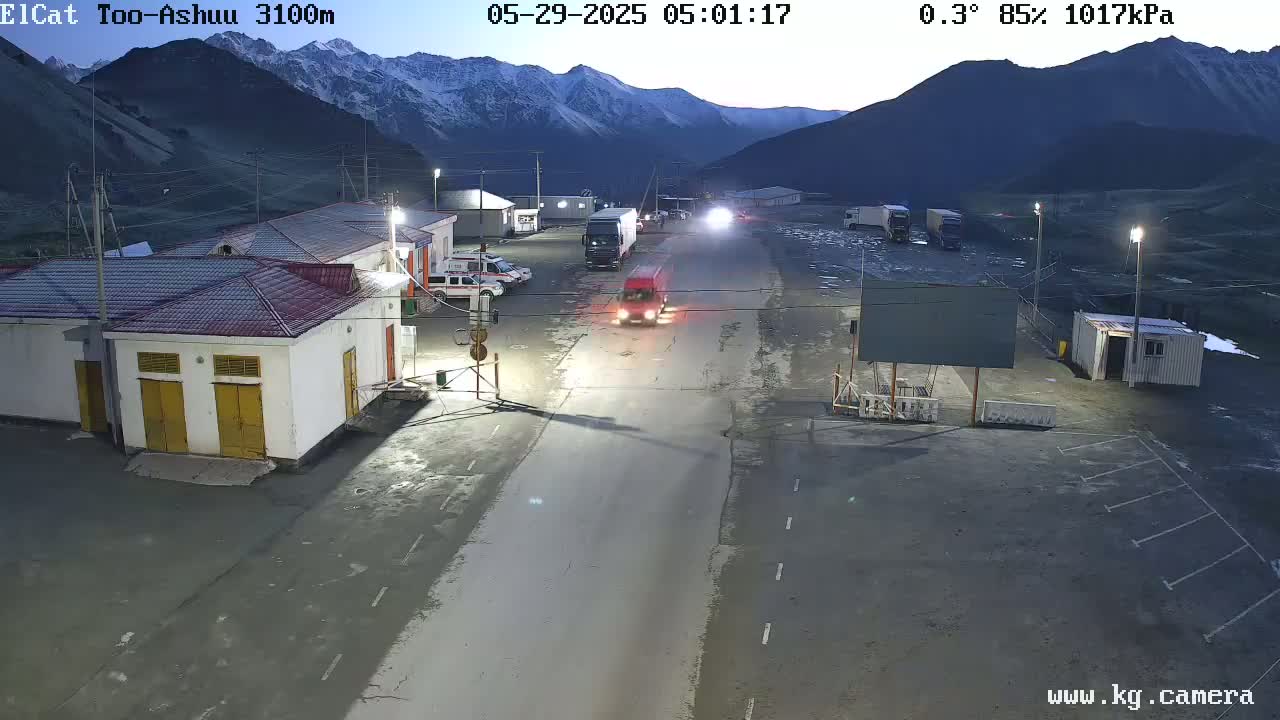 A road in a mountainous area at dawn, with several vehicles parked and one driving, near buildings and under a clear, cold sky (0.3°C, 85% humidity).