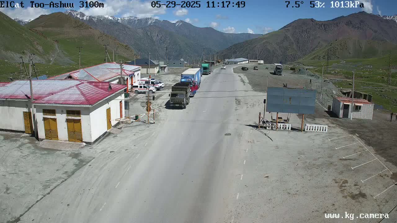 A highway winds through a mountainous region under a partly sunny sky, with several vehicles parked near buildings at a roadside checkpoint.