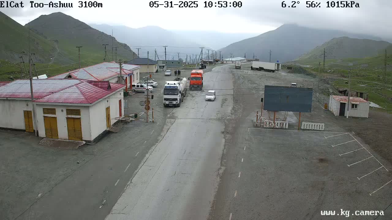 A road with several vehicles passes through a mountainous area on an overcast day, with buildings and a parking area visible to the side.