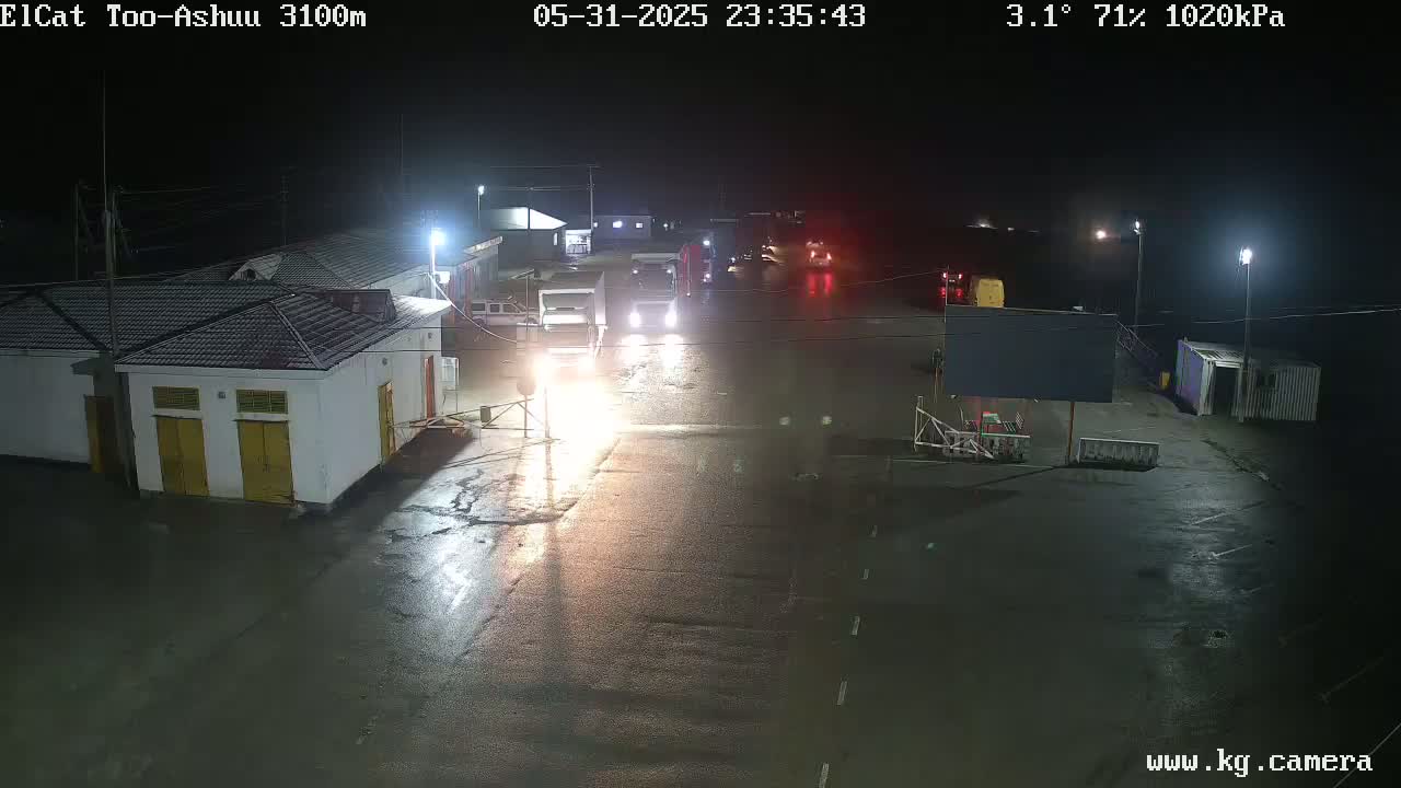 A wet, nighttime scene shows several vehicles at what appears to be a border crossing or checkpoint, with a building in the foreground and other structures in the background under a dark sky.