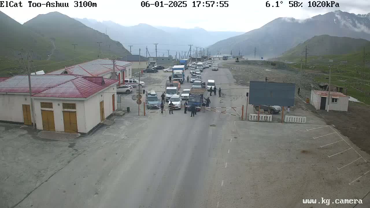 A line of vehicles is stopped at a border crossing in a mountainous area under cloudy skies.