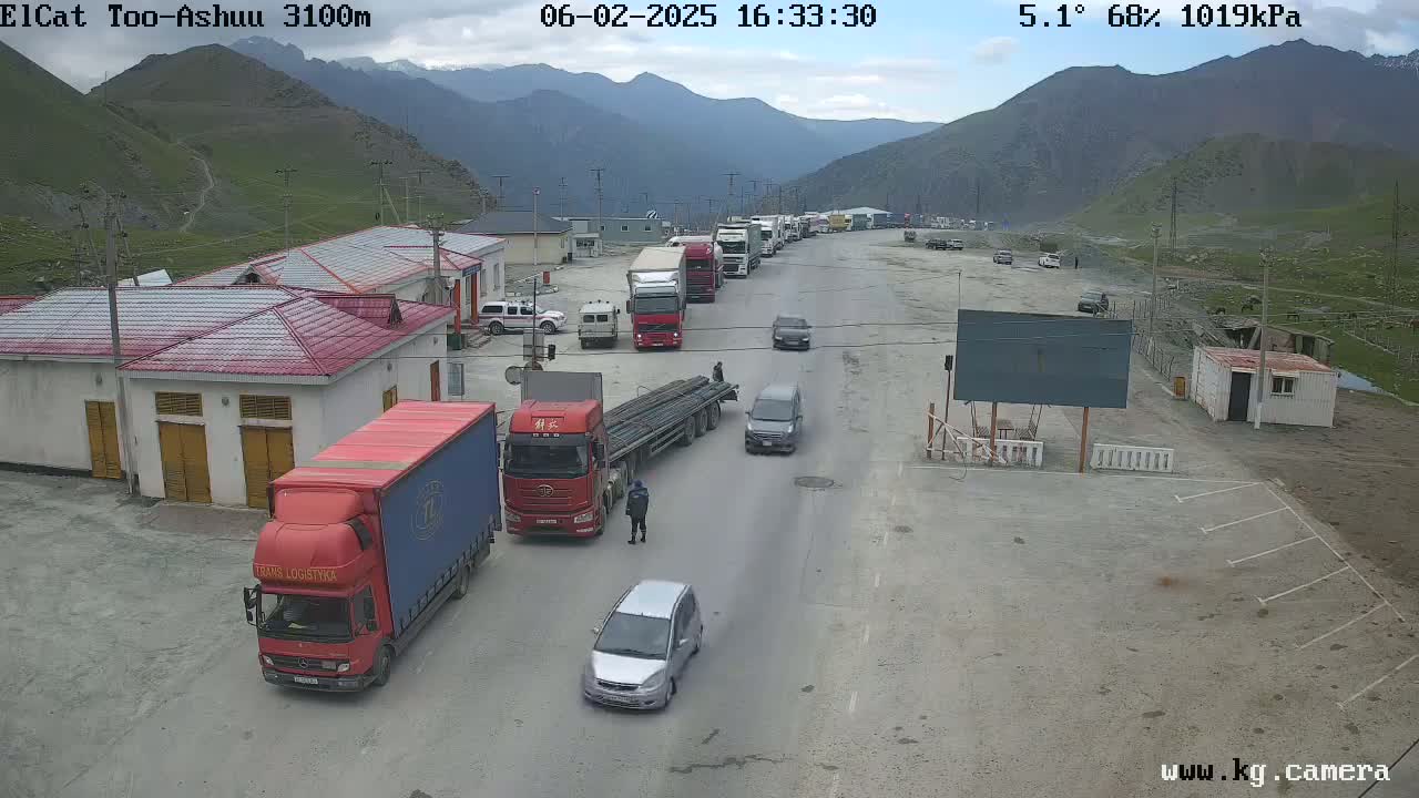 A line of trucks and cars is stopped at a mountain border crossing under a partly cloudy sky.