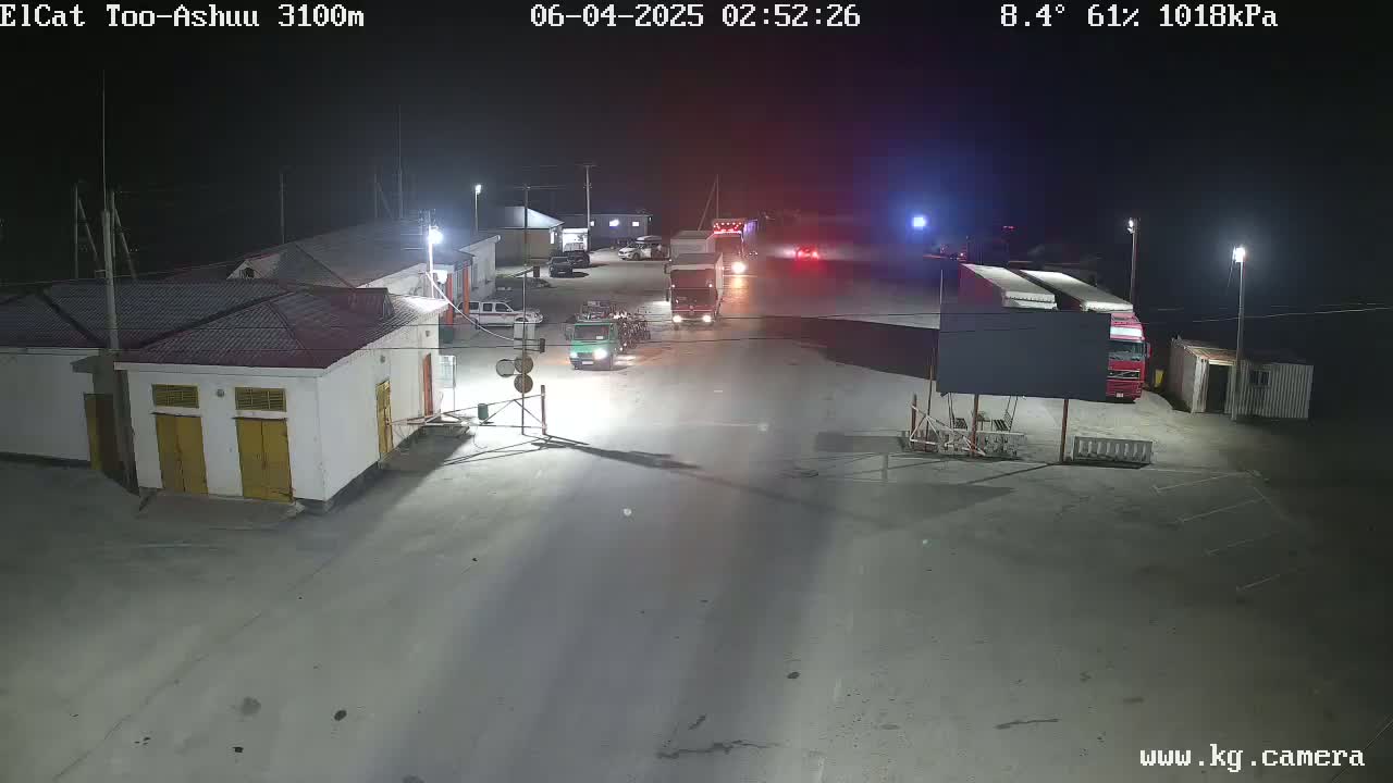 At night, several vehicles, including trucks, are parked and moving in a large, well-lit outdoor area near buildings.