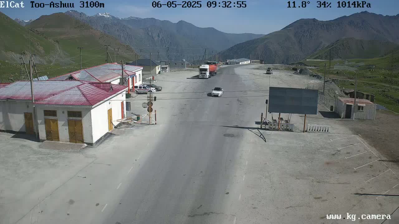 A highway checkpoint in a mountainous region on a sunny day shows several vehicles, including a truck and cars, near buildings, with clear skies and mountains in the background.