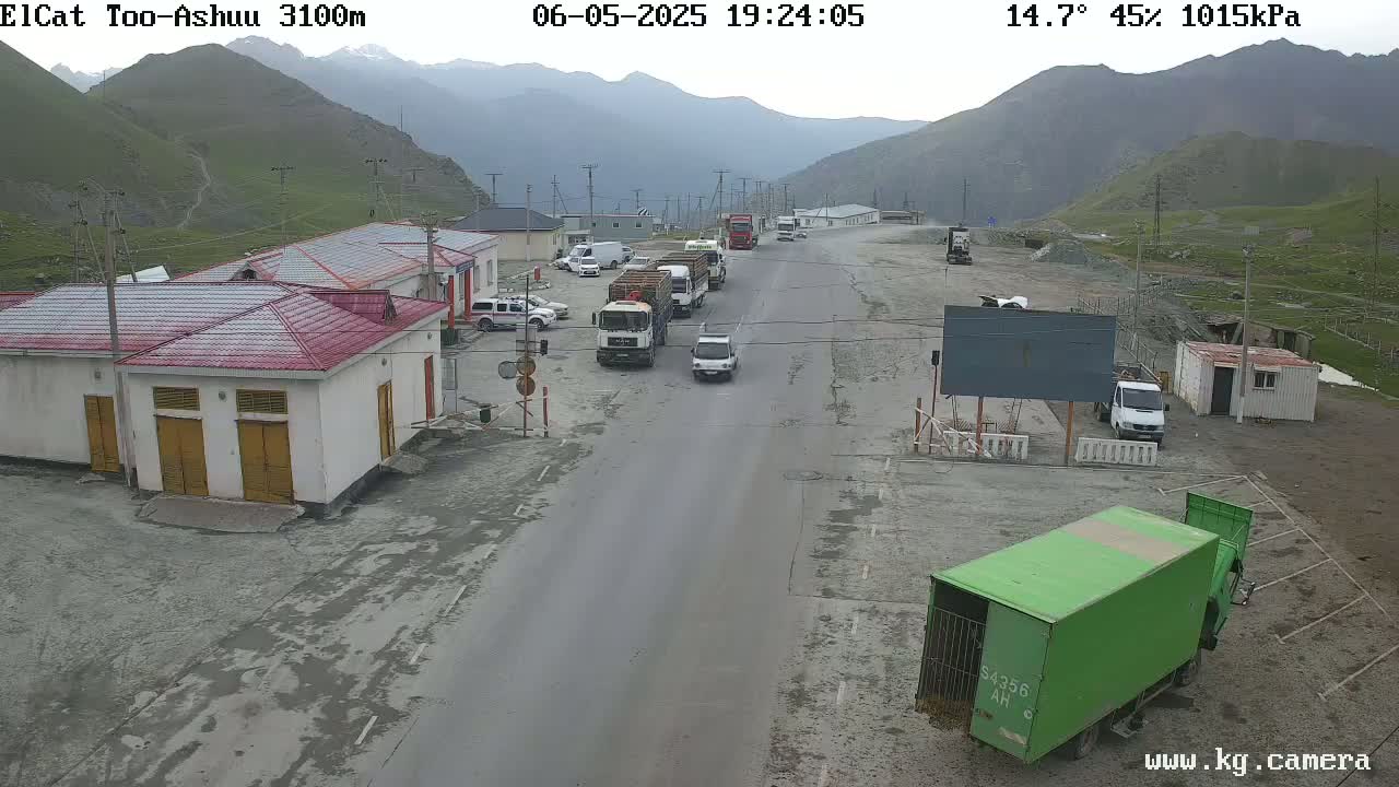 A mountain road scene shows several vehicles including trucks and cars parked and driving along a road in front of buildings, under an overcast sky.