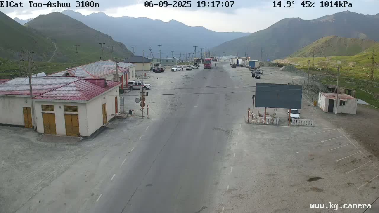 A mostly empty roadway and parking lot are situated in a mountainous area with overcast skies, a few vehicles are present.