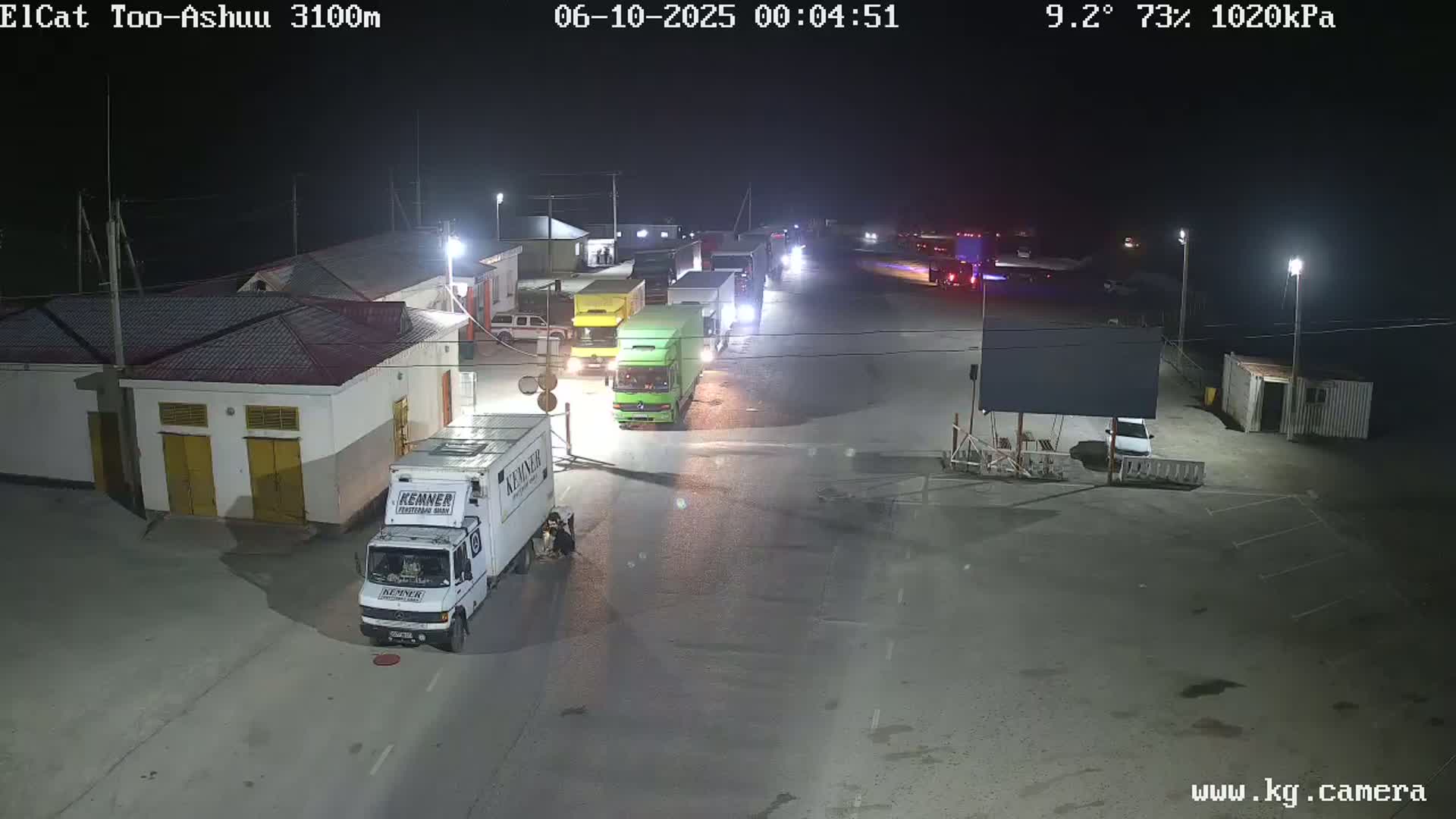 A nighttime view of a parking lot with several trucks and buildings, under a clear sky with a temperature of 9.2 degrees and 73% humidity.