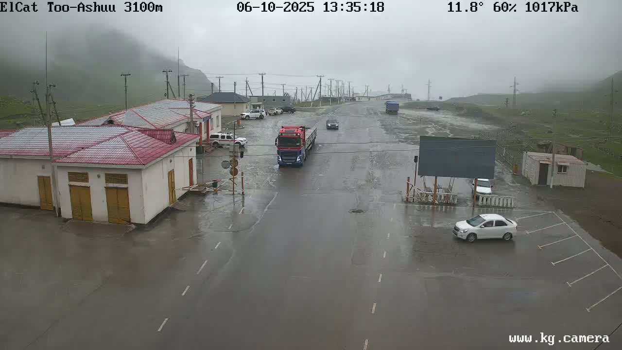 A wet road at a border crossing station in mountainous terrain shows several vehicles, including a semi-truck, parked and driving in the rain.