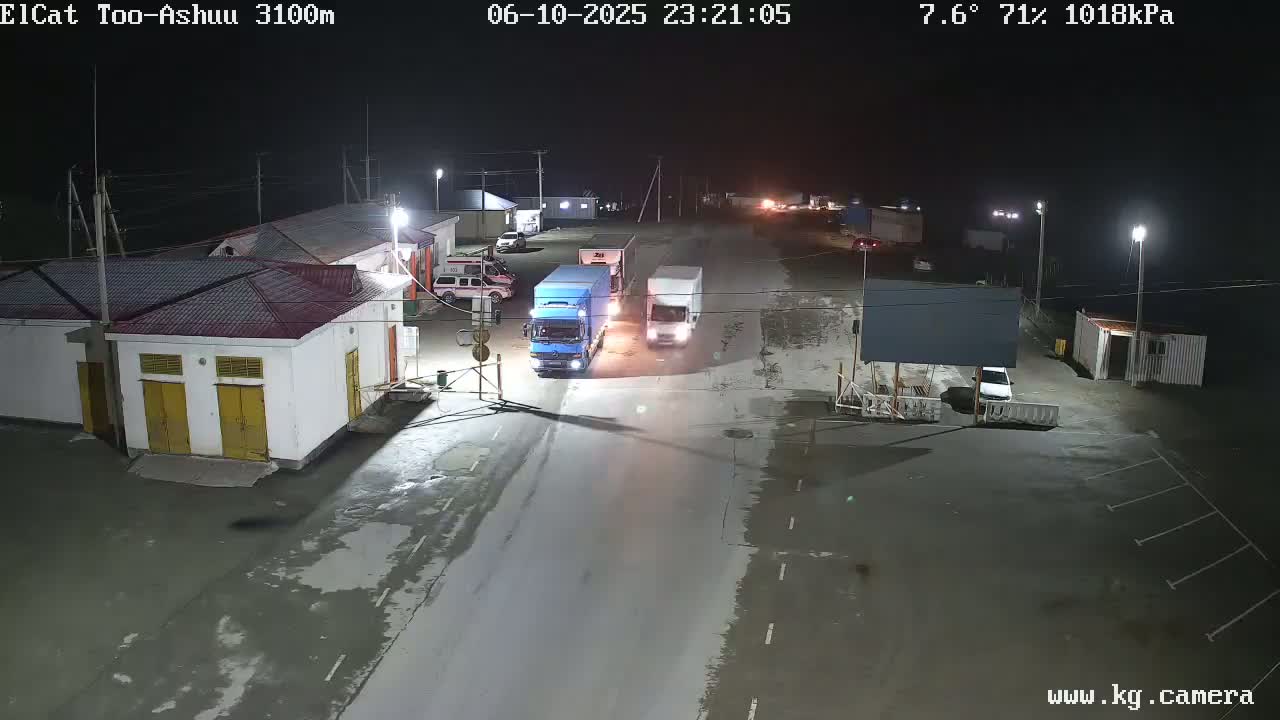 At night, several vehicles, including trucks and a car, are parked or driving in a mostly empty, paved lot near a building complex under a clear, cool sky.