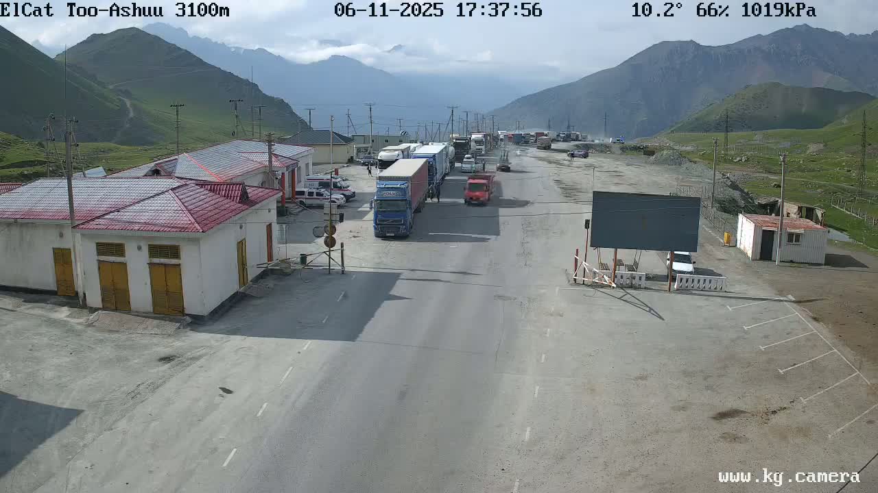 A mountain road with several trucks and cars stopped at a checkpoint under partly cloudy skies.