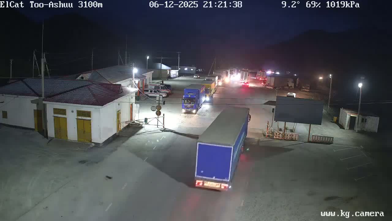 At night, under a partly cloudy sky with a temperature of 9.2 degrees Celsius and 69% humidity, several trucks and cars are parked in a brightly lit outdoor area, near some buildings.
