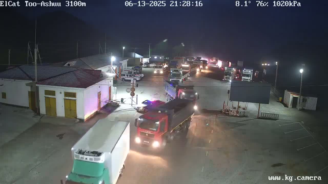 At night, under a mostly clear sky with a temperature of 8.1°C and 76% humidity, numerous trucks are parked and driving in a large paved area next to a building complex.
