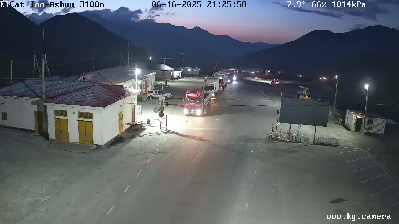 Several trucks are parked in a dimly lit area at night, near buildings against a backdrop of dark mountains under a twilight sky.