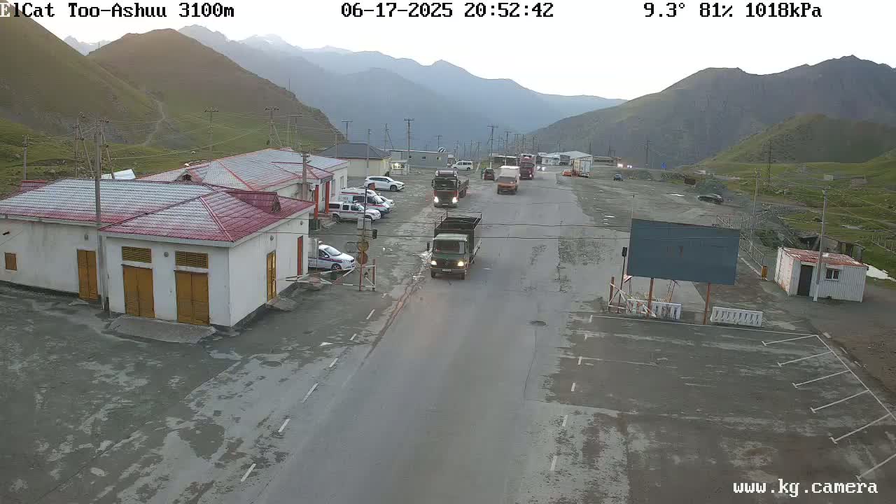 Several trucks and cars are parked and driving on a road in a mountainous area under a partly cloudy sky.